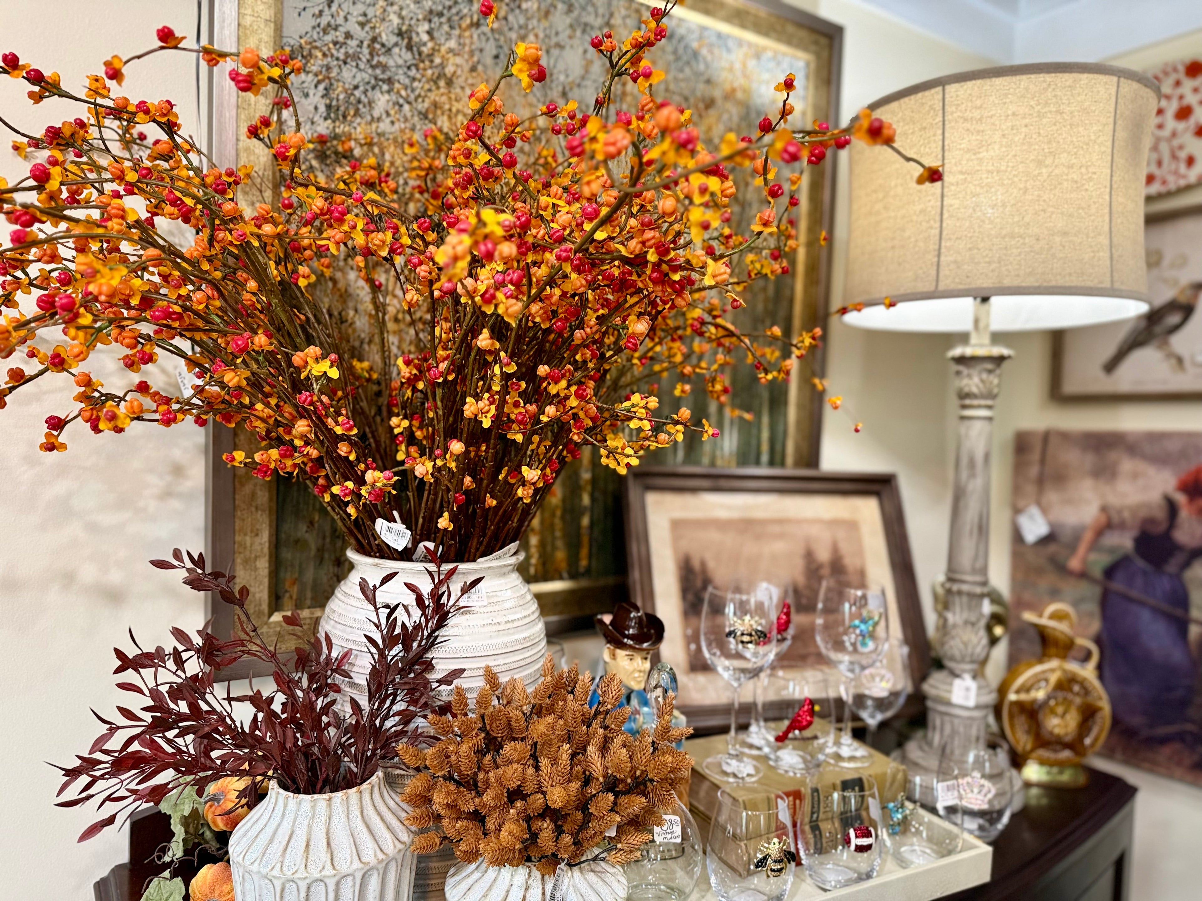 A large artificial autumn floral arrangement in a white ribbed vase, displayed on a table alongside a table lamp, wine glasses, and framed artwork in an indoor setting.