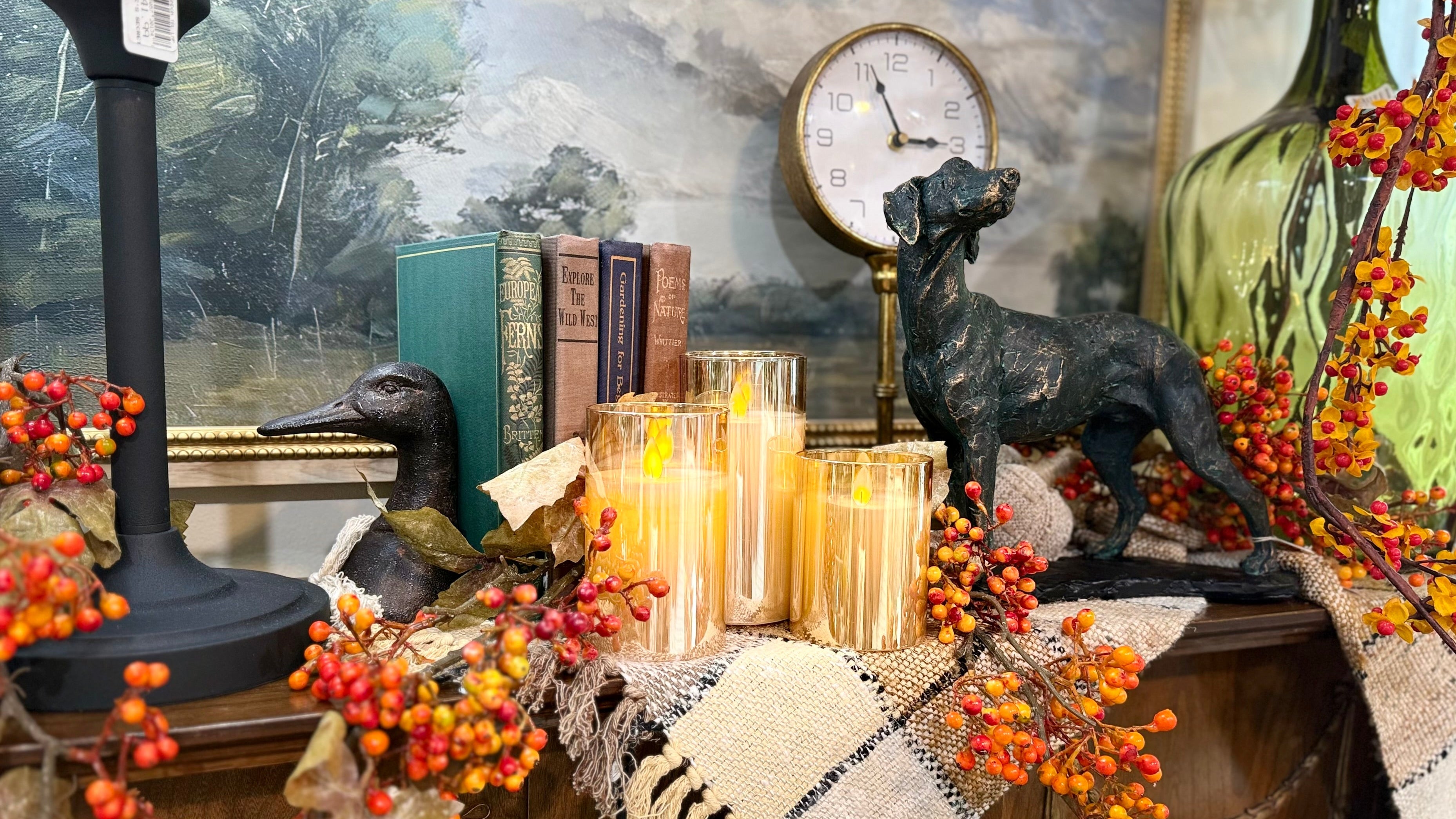 A styled autumn-themed tabletop display featuring a black dog statue, a duck figurine, a black candlestick base, several lit glass candle holders, vintage-style books, and orange berry garlands against a scenic landscape wall mural.