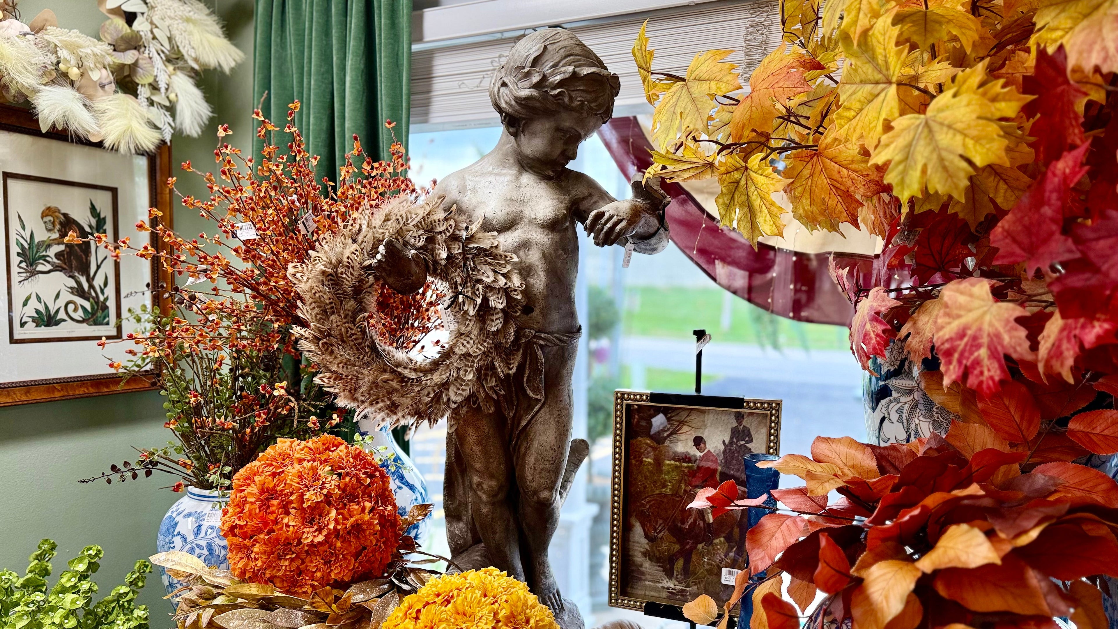 A styled indoor arrangement featuring a central classical-style statue holding a wreath, surrounded by autumn artificial foliage, orange and yellow flower balls, pumpkins, framed art, and blue-and-white ceramic vases.