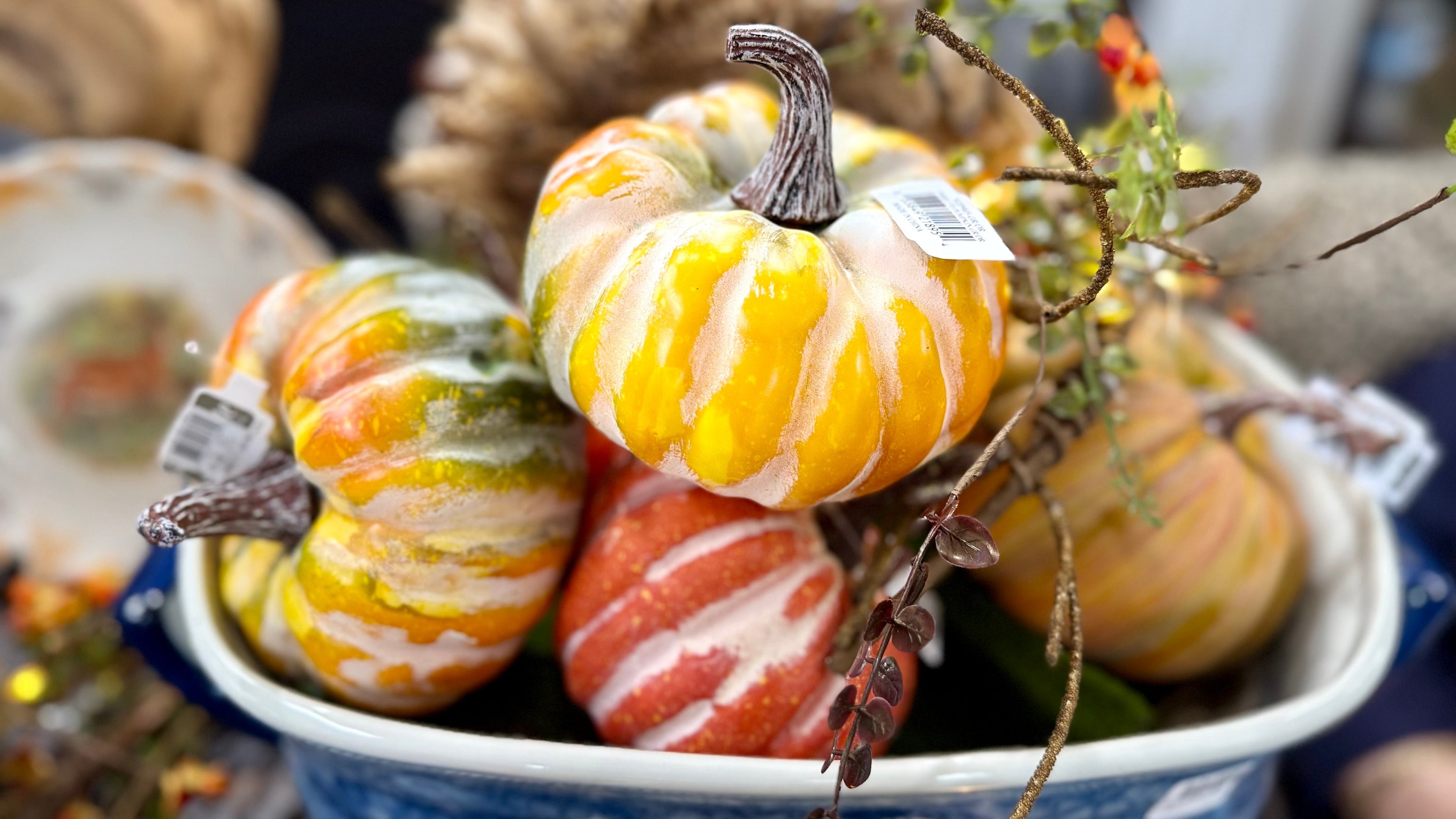A decorative arrangement of small striped pumpkins and gourds in a blue-and-white ceramic bowl, styled with dried grasses and branches in a real indoor setting.
