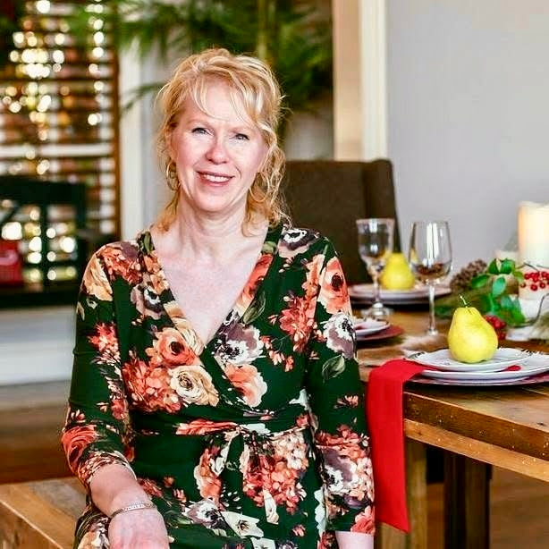 A smiling blonde woman wearing a dark green floral wrap dress with orange, red, and cream flowers, seated at a wooden dining table set with wine glasses, plates, and a pear in a warmly lit indoor setting.