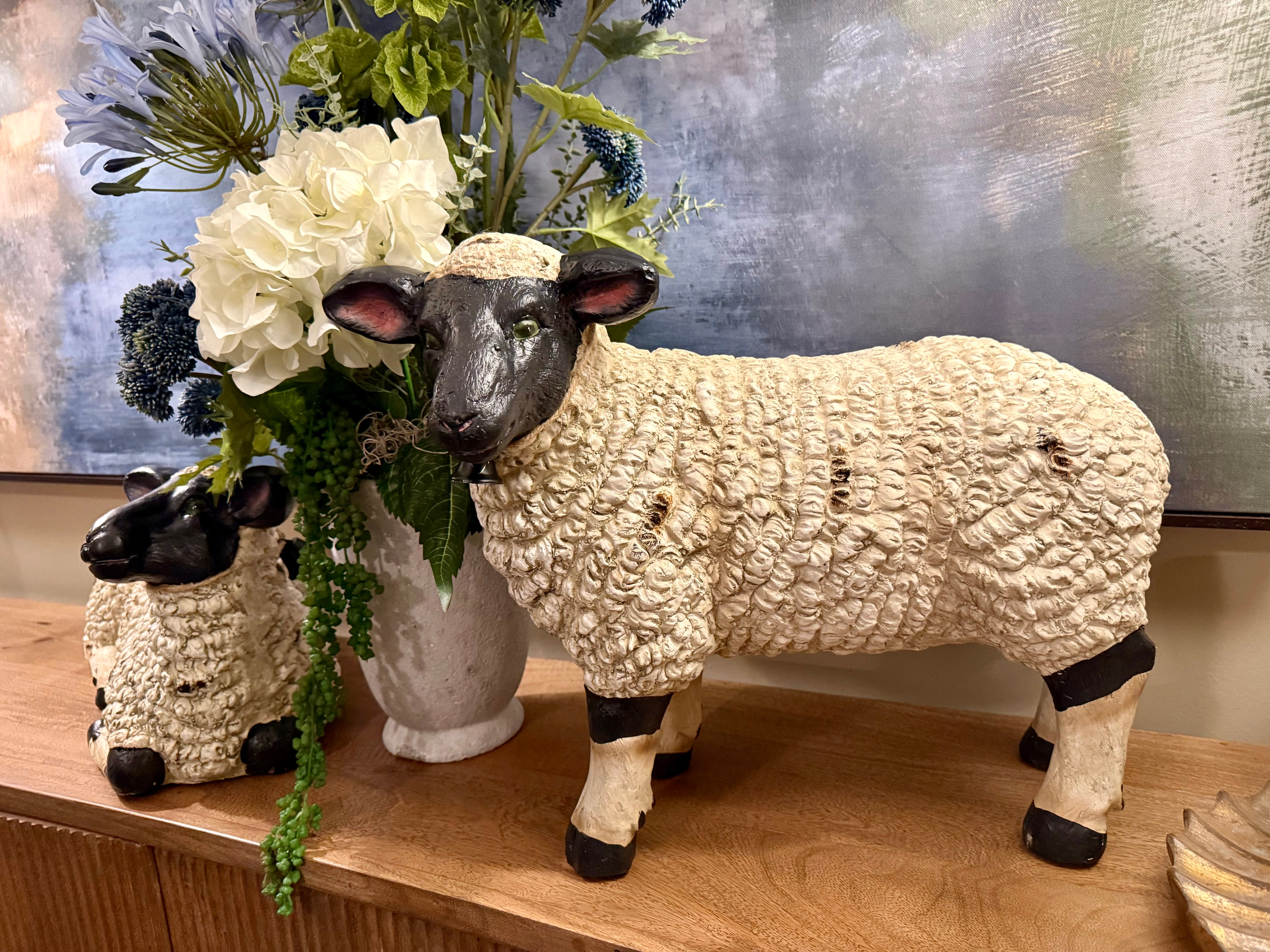 Two textured sheep figurines, one large and one small, styled with a faux flower arrangement on a wooden shelf in front of a blue-gray abstract painting.